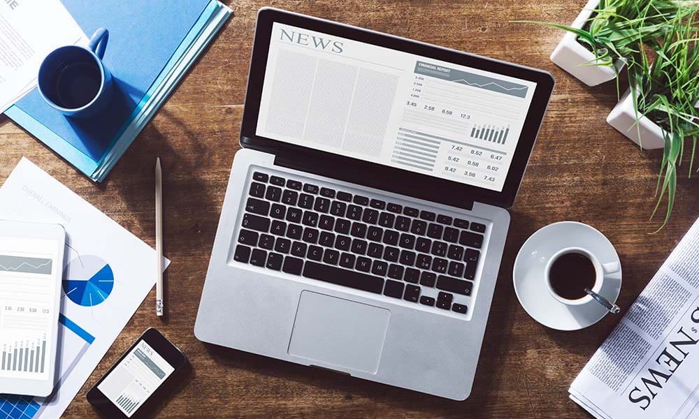 Magenta Blog Sample Post - View of a Laptop with the News Sitting on Top of a Wooden Desk Next to a Cup of Coffee and Paperwork